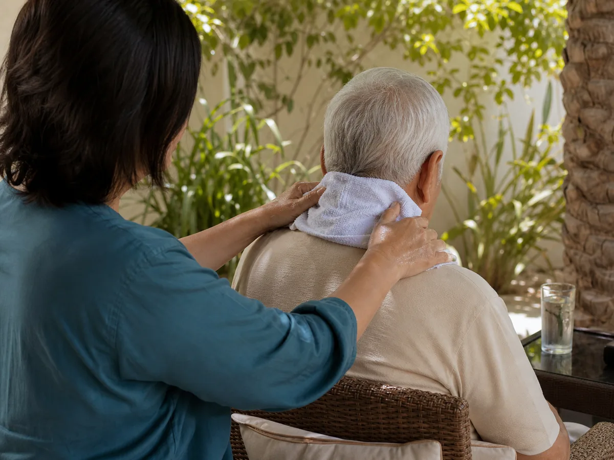 Person applying a cold, wet towel to the back of an elderly relative's neck in a shaded Dubai garden, midday light, editorial documentary style